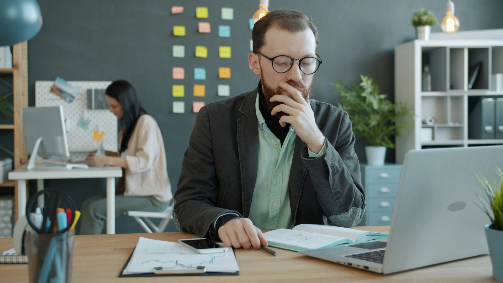 A thoughtful man pauses beside a laptop, notebook, and papers at an office desk.